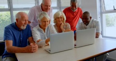 Huzurevinde dizüstü bilgisayar kullanan aktif karışık ırk lı yaşlıların ön görünümü. Masada oturuyorlar. 