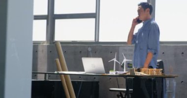 Modern bir ofiste cep telefonu yla konuşan Asyalı erkek mimarın ön görünümü. Dizüstü bilgisayar kullanıyor. 