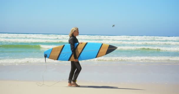 活跃高级白种女性冲浪者在阳光下在海滩上散步的侧视图 她拿着冲浪板4K — 图库视频影像