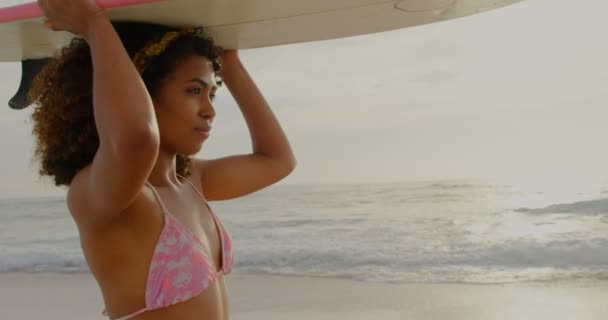 ビーチで彼女の頭の上にサーフボードを運ぶアフリカ系アメリカ人の女性サーファーの側面図 彼女はビーチ4Kで走っています — ストック動画