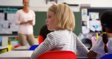 Mutlu Beyaz kız öğrencinin sınıfta kameraya bakabakan arka görüntüsü. Arka planda kadın öğretmen öğretim 