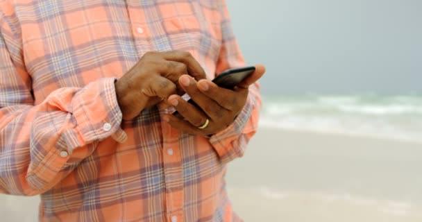 ビーチで携帯電話を使用してアクティブなシニアアフリカ系アメリカ人の男性の中間セクション 彼はテキスト4Kをテキストメッセージです — ストック動画