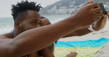 Sahilde cep telefonuyla selfie çeken Afrikalı-Amerikalı çiftin ön görünümü. Onlar plajda 4k rahatlatıcı