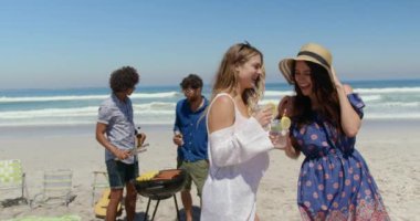 Erkekler güneşli bir günde arka planda barbekü yemek pişirirken plajda birbirleriyle konuşurken içecekler ile genç Kafkas kadınların ön görünümü 4k