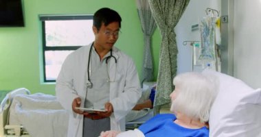 Asyalı erkek doktorun hastanedeki koğuşta aktif kıdemli hastayla etkileşime girmesi. Elinde dijital tablet 4k var.