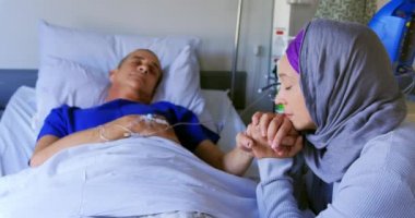 Hastanenin koğuşunda erkek hastanın elini tutan Asyalı kadının yan görüntüsü. 4K numaralı koğuşta uyuyan erkek hasta.
