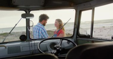 Sahilde camper minibüsün yakınında birbirlerini kucaklayan Kafkas çiftinin yan görünümü. Onlar 4k gülümsüyor