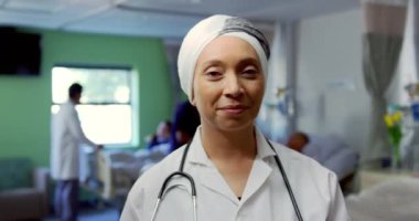 Hastanenin koğuşunda dikilen melez kadın doktorun ön görüntüsü. Gülümsüyor ve kamera 4k 'ye bakıyor.