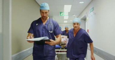 Hastanenin koridorunda acil sedye yatağı iten çok ırklı tıbbi ekiplerin ön görüntüsü. Erkek doktor not defterine 4K yazıyor.