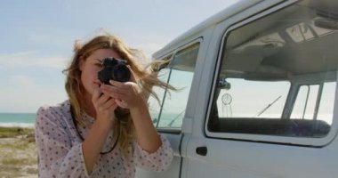 Güneşli bir günde plajda karavan ın yanında dururken dijital kamera ile fotoğraf çeken genç Kafkas kadının ön görünümü 4k