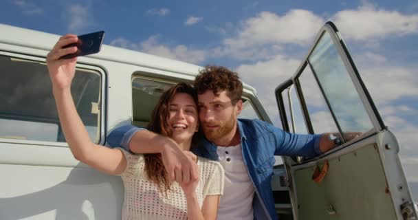前视图年轻的高加索夫妇采取自拍与手机附近的露营车在海滩 而微笑和摆出4K — 图库视频影像