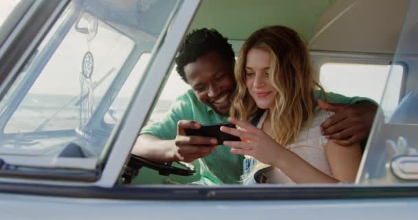 ビーチでキャンピングカーで携帯電話を使用して多民族カップルの正面図 彼らは微笑んで楽しい4Kを持っています — ストック動画
