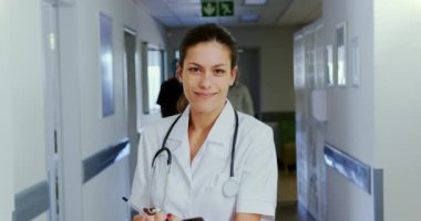 Hastanenin koridorunda dikilen beyaz kadın doktorun ön görüntüsü. Gülümsüyor ve kamera 4k 'ye bakıyor.