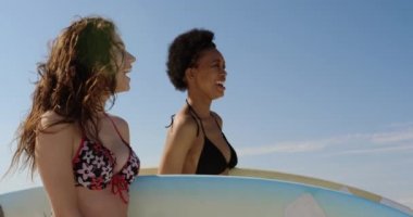 Güneşli 4k plajda sörf tahtası ile ayakta iken konuşan ve gülümseyen genç çok etnik kadın arkadaşlar Yan görünümü