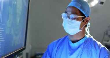 Hastanede ameliyat hanesinde röntgen raporunu inceleyen Asyalı erkek cerrahın ön görünümü. Monitör 4k'ye bakıyor.