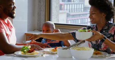 Afro-Amerikan ailenin rahat bir evde yemek masasında yemek yemesi ön planda. Annem yemek servisi yapıyor. 4k