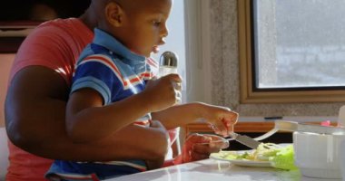 Afro-Amerikan baba ve oğlunun evde yemek masasında yemek yeme yan görünümü. Oğlum salataya tuz koyuyor. 4k
