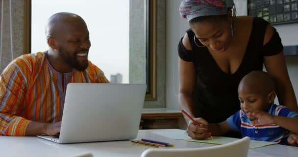 アフリカ系アメリカ人の母と息子が自宅のテーブルの上の紙にスケッチを描く正面図 父は微笑んで見ている — ストック動画