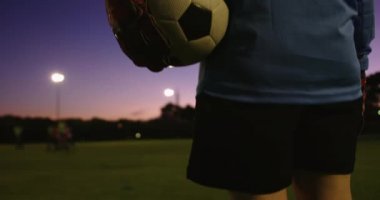 Kadın futbolcunun arkadan görüntüsü, gece futbol sahasında kaleci eldiveni giyerken topu tutuyor. 4k