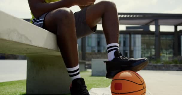 バスケットボールコートで携帯電話を使用してアフリカ系アメリカ人バスケットボール選手の正面図 彼は水4Kを飲んでいます — ストック動画