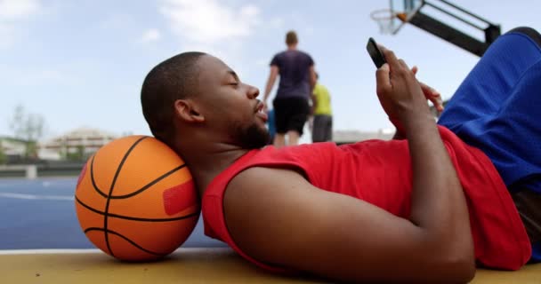 バスケットボールコートで携帯電話で話しているアフリカ系アメリカ人バスケットボール選手の側面図 彼は法廷4Kで地面に横たわっている — ストック動画