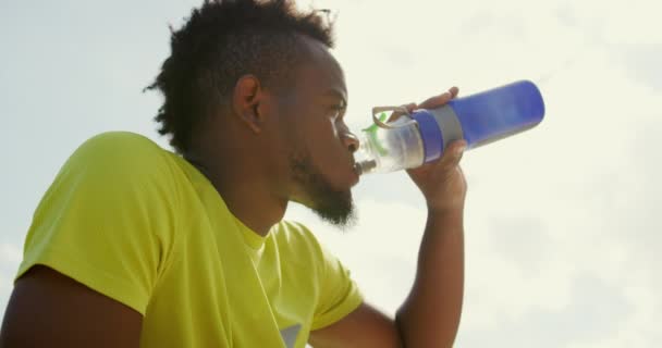 バスケットボールコートで携帯電話を使用してアフリカ系アメリカ人バスケットボール選手の低角度ビュー 彼は水4Kを飲んでいます — ストック動画