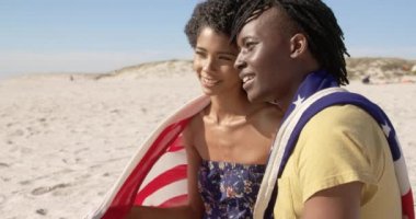 Sahilde birlikte oturan Amerikan bayrağına sarılı Afrikalı Amerikalı çiftin yan görünümü. Birbirleriyle etkileşime giriyorlar 4k