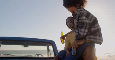 Sahilde bir kamyonetin üzerinde oturan erkek kucağında Afrikalı Amerikalı kadın. Bira 4k içiyorlar.