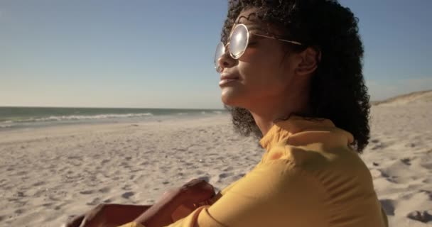 戴着太阳镜的非洲裔美国妇女在海滩上放松的侧视图 她看着远方4K — 图库视频影像