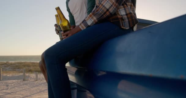 低角度的非洲裔美国人夫妇与啤酒瓶坐在小卡车上在海滩 他们微笑着说着4K — 图库视频影像
