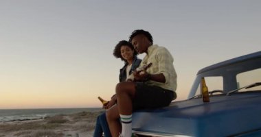 Gitar çalan Afro-Amerikan erkeğin yan görünümü ve kamyonet kaputunda şarkı söyleyen kadın. Bira 4k'ı yediyorlar.