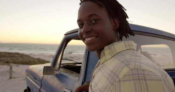 ビーチでピックアップトラックの近くにカメラを見ているアフリカ系アメリカ人の男性の側面図 彼は4Kを微笑んでいる — ストック動画