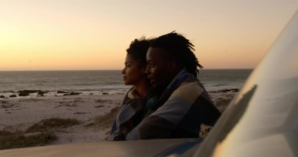 日没時にビーチでピックアップトラックの近くに毛布に包まれたアフリカ系アメリカ人カップルの側面図 4Kを見つめている — ストック動画
