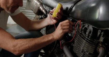 Beyaz erkek tamircinin yan görünümü tamir garajında motosiklet bataryalarını kontrol ediyor. Multimetre 4k kullanıyor.