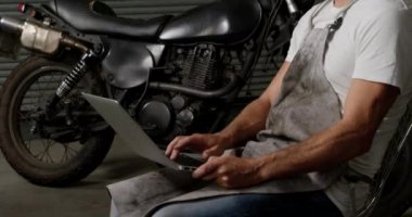 Motosiklet tamir garajında dizüstü bilgisayar kullanarak Kafkas erkek mekanik yan görünümü. O sandalyede 4k oturarak