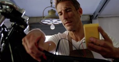 Beyaz erkek tamircinin ön görünümü tamir garajında motosiklet bataryalarını kontrol ediyor. Multimetre 4k kullanıyor.