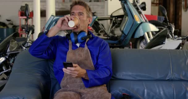 在摩托车修理厂使用手机的白种男性机械师的正面视图 他正在喝咖啡4K — 图库视频影像
