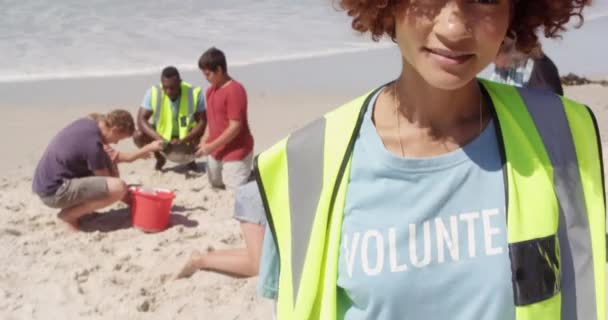 ビーチでカメラを見るアフリカ系アメリカ人女性ボランティアのフロントビュー 多様なボランティアを背景に笑顔でいる — ストック動画