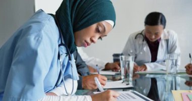 Masa yazıları yazan tesettürlü genç ve melez bir kadın doktorun el kamerasıyla çekilmiş fotoğrafı. Durdu, kameraya döndü ve gülümsedi. Arka planda çalışan iki genç doktor daha var. Ön plana odaklan. 4k