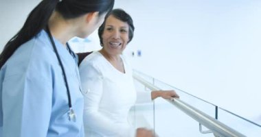 Hastane koridorunda tırabzan tutan yaşlı melez bir hastanın yan görüntüsüne dost canlısı genç bir bayan doktor yardım ediyor.