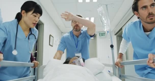 緊急時に病院の廊下の下のベッドで患者を押す3人の多民族病院労働者の逆追跡ショットを閉じる — ストック動画