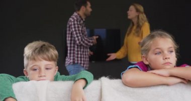 Beyaz çocukların kanepeye yaslanıp ebeveynleri evde arka planda tartışırken ön görünümü. Onlar üzgün ve üzgün 4k