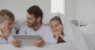 Beyaz baba ve çocukların ön görünümü evde yatakta battaniye altında dijital tablet kullanarak. Onlar gülümsüyor ve mutlu 4k