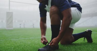 Stadyumda ayakkabı bağı bağlayan Afrikalı Amerikalı rugby oyuncusunun yakın çekimi. Takım arkadaşları arka planda 4k oynuyorlar