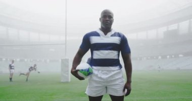 Afrika kökenli Amerikalı rugby oyuncusu stadyumda ragbi topuyla dikiliyor. 4K kamerasına bakıyor.