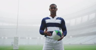 Afrika kökenli Amerikalı rugby oyuncusu stadyumda ragbi topuyla dikiliyor. 4K kamerasına bakıyor.