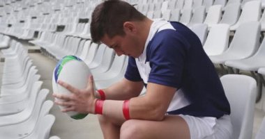 Kafkas erkek rugby oyuncusu stadyumda ragbi topuyla oturuyor. Morali bozuk ve başka tarafa bakıyor.  