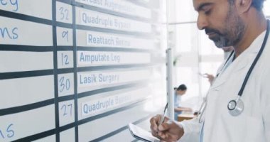 Orta yaşlı, melez bir erkek doktorun beyaz bir tahtadaki bilgileri kontrol edip notlar alması el kamerasıyla yakından görülüyor. 