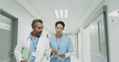 Son sınıf melez erkek doktor ve beyaz kadın doktorun tablet bilgisayarla hastane koridorunda yürürken görüntülerini ters çevirin ve konuşun. 