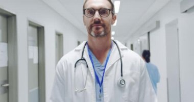 Hastanenin koridorunda kameraya gülümseyen orta yaşlı beyaz erkek doktorun yavaş çekim kafası ve omuzları. Hastane personeli geçiyor.. 